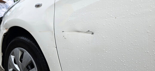 The left front door of the car has a deep scratch from an accident while it was raining. The white car has water droplets on door. Damage occurred after the accident. or driving and hitting house door