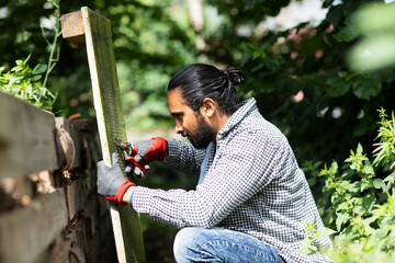 A man kneels outdoors, measuring a wooden plank with a tape measure in a lush garden. Germany