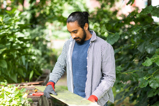 A man holding a wooden board in a green garden setting with tools nearby. Germany