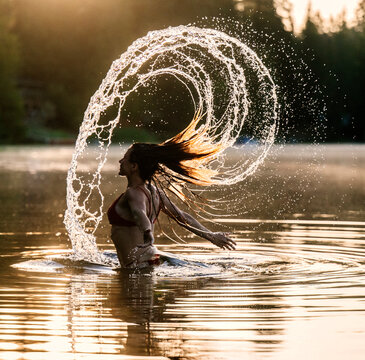 Woman in water flips hair creating arc of droplets at sunset in serene lake setting. WA, USA