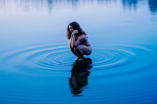 Woman crouching in clear blue water creating ripples around her reflection. WA, USA