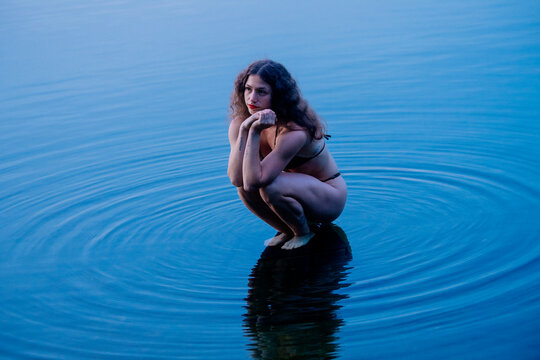 Woman crouching in clear blue water creating ripples around her reflection. WA, USA