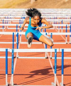 Athlete in blue jumpsuit clears hurdle on outdoor track with determination and focus. USA