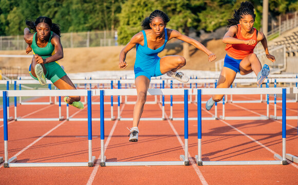 Three athletes in colorful outfits hurdle along a red track under a sunny sky. USA
