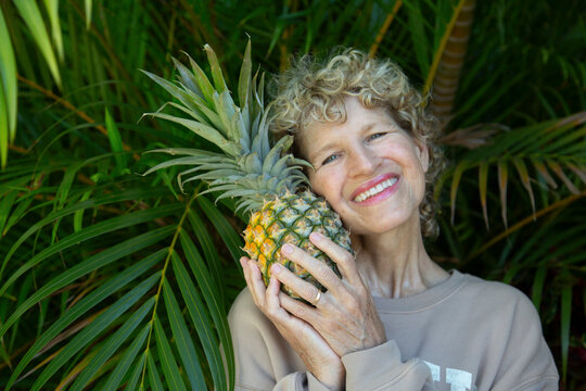 Smiling woman holding a pineapple in front of lush green palm leaves. Kepaniwai Park, Wailuku, Hawaii