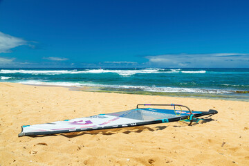 Windsurfing board rests on a sandy beach under a clear blue sky with ocean waves in the background. Hookipa beach park, Maui, Hawaii