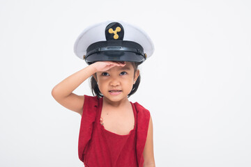A delighted young Asian girl saluting with one hand, smiling joyfully and wearing a pilot cap, exuding charm and pride. Isolated on a white background.