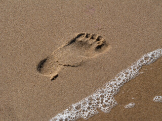 Human footprint in wet sand near the sea with a gentle wave approaching, symbolizing travel, relaxation, and fleeting moments
