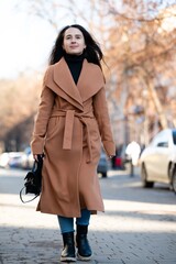 Fototapeta premium Walking confidently on a sunny street, a young woman in a stylish coat and jeans embraces the warmth of the day as autumn leaves fall around her