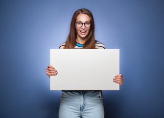 Fototapeta premium Promoting women holding white board, isolated on blue background. Amused girl showing blank empty paper billboard with blank space for text.