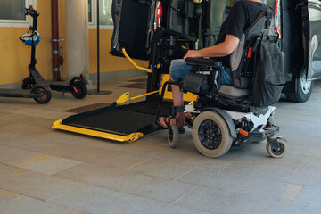 Person with disability uses a powered wheelchair and lift to board an accessible vehicle at a transport hub during the day.