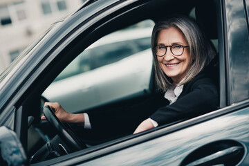Elegant mature businesswoman driving a stylish car while smiling brightly in an urban city environment during daytime © deagreez
