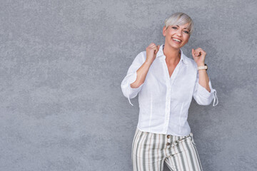 Amused beautiful adult woman with closed eyes and clenched fists, against gray background - wall. Yes concept. Amusing middle age woman celebrates success