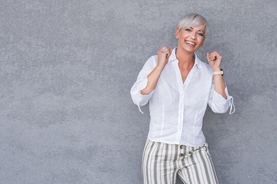 Amused beautiful adult woman with closed eyes and clenched fists, against gray background - wall. Yes concept. Amusing middle age woman celebrates success