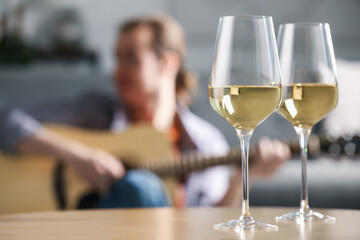 Glasses with white wine on table and man playing guitar indoors, selective focus