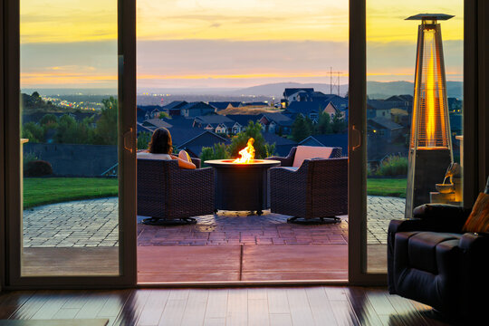 A young woman relaxes on a patio chair with a flaming fire pit nearby as she enjoys the view of the valley and illuminated city at Golden Hour sunset.