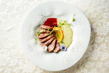 Gourmet duck breast slices with orange, microgreens, and edible flowers, plated with red and green sauces on a white dish over floral tablecloth