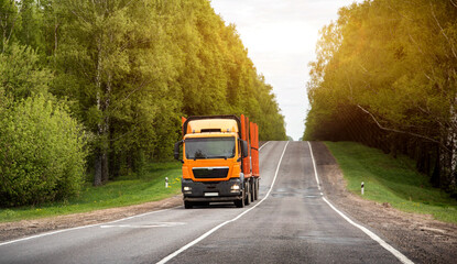 Fototapeta premium Modern new orange timber truck with semi-trailer on country road against forest and cloudy sky background. Copy space for text, industry