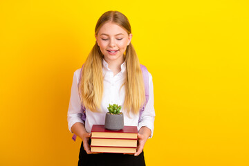 Cheerful schoolgirl in a white shirt holding books and a potted plant against a vibrant yellow background