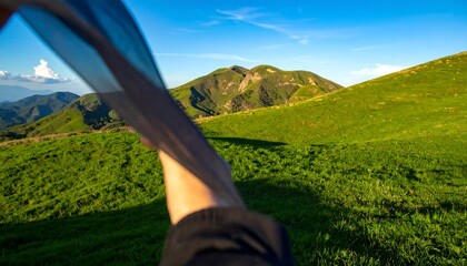 A hand holds a sheer fabric, capturing a vista of rolling hills and distant mountains under a vibrant blue sky.