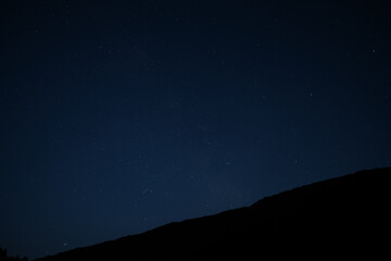 Obraz premium Starry night sky above mountain silhouette, with faint meteors and constellations visible in deep blue. View of the Milky Way from the northern hemisphere.