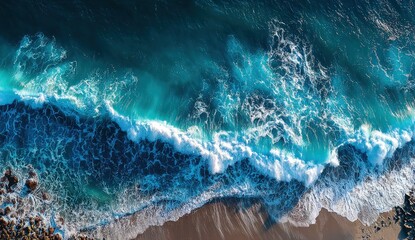 An aerial perspective reveals ocean waves crashing onto a sandy beach, with varying shades of blue and white foam