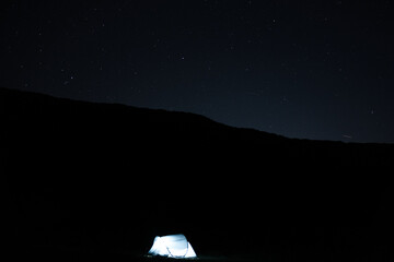 Illuminated camping tent under a starry night sky with visible meteors, creating a peaceful outdoor...