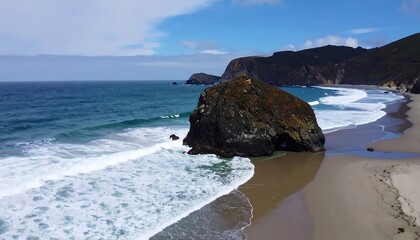 Expansive ocean view of a dramatic coastline with waves crashing against a large, dark rock formation on a sandy beach.