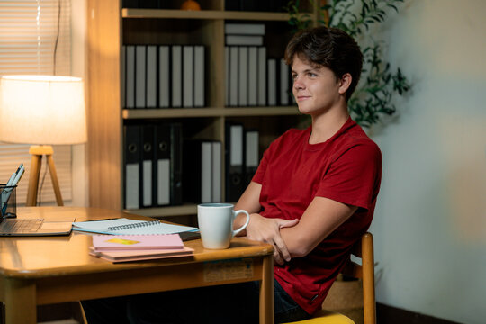 Teenager taking a break from working or studying, sitting at desk with laptop, notebooks and coffee mug, looking away