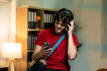 Teenage male listening to music, wearing wireless headphones, scrolling smartphone near bright window with minimalist room interior