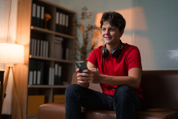 Obraz premium Young man relaxing on leather couch, wearing headphones around neck, browsing smartphone with cheerful smile in home living room