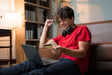 Happy teenager making online payment using credit card and laptop, celebrating successful purchase from home