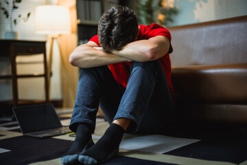 Depressed young male sitting on living room floor, head bowed between knees, expressing emotional distress and mental struggle