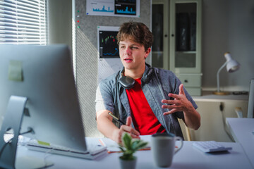 Young man engaging in a video call while taking notes on a notepad, working comfortably from his modern home office during the day