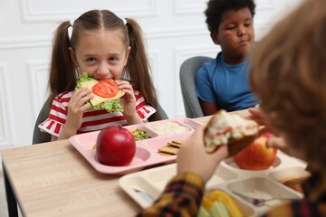 Little kids eating lunch at wooden table in school canteen