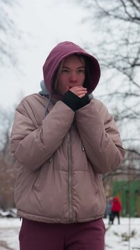 Calm woman in winter outfit strolling alone through a snow-covered park, bringing hands close to mouth for warmth, bare trees, scattered snow, and blurred figures in the background