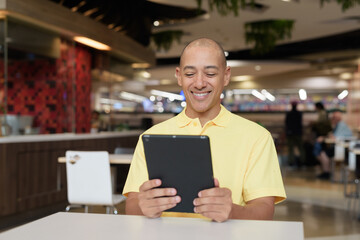 Middle-aged bald Hispanic man using tablet computer while dining at food court restaurant