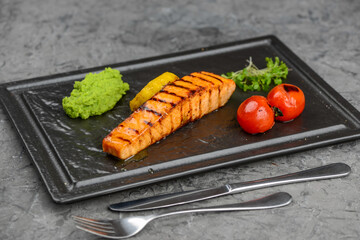 Grilled salmon fillet in a black plate with tomatoes on a gray background