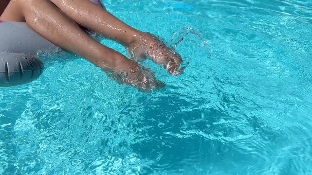 Close-up of a girl's feet splashing in the bright blue water of a swimming pool while relaxing on an inflatable float. Summer vacation fun. Holiday lifestyle