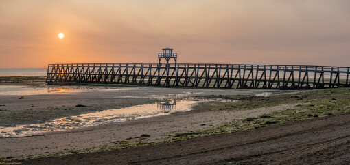 Luc-Sur-Mer, France - 08 10 2025: Panoramic view of the wooden pier in front of the sea named Fisherman's Pier at sunrise with the sun rising and Le Havre far away behind