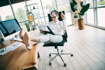 Confident businesswoman relaxing in a modern office setting, reviewing documents at her desk with professionalism and style