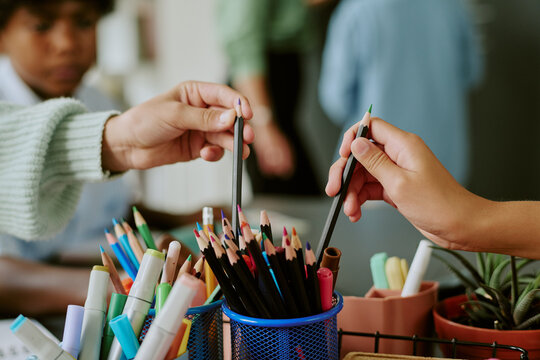 Caucasian child and Black child reaching for colored pencils from container, hands meeting above assortment of drawing supplies, blurred classmates visible in background, creative activity