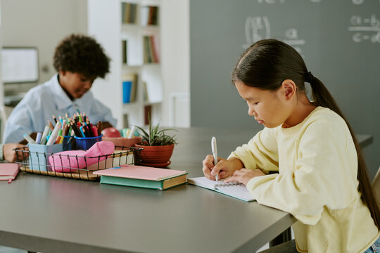 Asian girl writing in notebook at desk while Black boy sitting in background drawing with colored pencils in classroom setting, both children focused on schoolwork, supplies visible on table