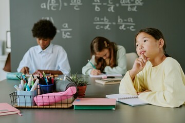 Asian girl sitting at desk thinking while Black teenager and Caucasian girl writing in notebooks in classroom with math equations on chalkboard in background, school supplies on table