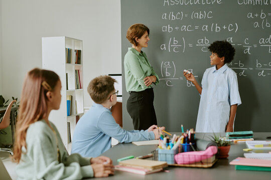 Caucasian female teacher interacting with Black teenage boy solving math equations on chalkboard, while two Caucasian teenagers and one Caucasian girl sitting at table observing classroom activity