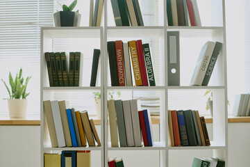 Bookshelf standing in bright room holding textbooks and novels, including mathematics, chemistry, algebra, coding science, with green plants placed on top and in background
