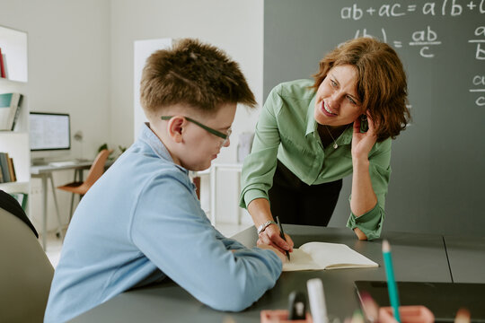 Caucasian middle aged woman leaning over desk assisting Caucasian teenage boy with schoolwork in classroom setting, both focused on open notebook, chalkboard with math equations in background