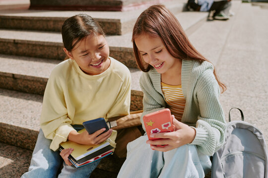 Two teenage girls, one Asian and one Caucasian, sitting on outdoor steps smiling while holding smartphones and books, engaging in conversation and sharing content together