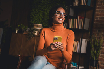Young woman in casual turtleneck sweater sitting indoors, holding smartphone, smiling confidently.