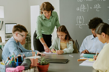 Middle aged Caucasian woman assisting diverse group of children working together at classroom table, students focusing on assignments while teacher providing guidance near chalkboard